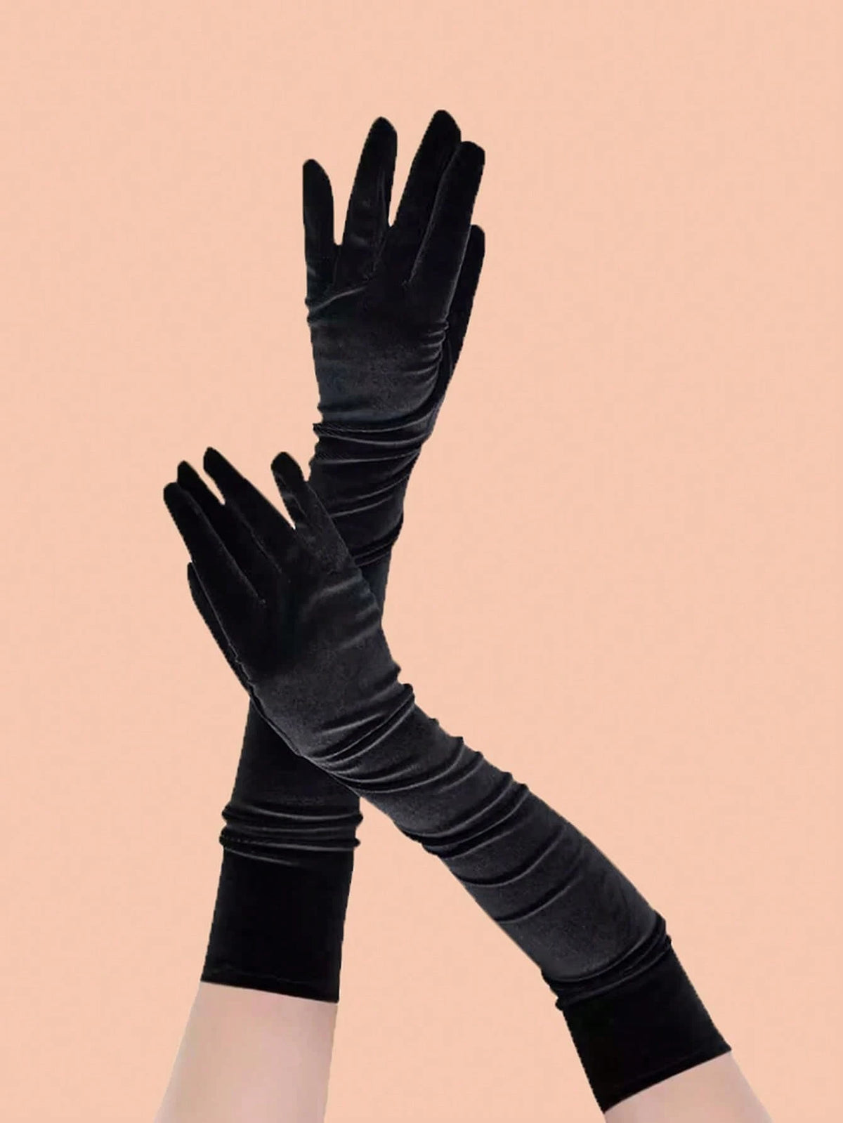 A pair of long black velvet opera gloves worn on hands, crossed over each other against a solid peach-pink background.