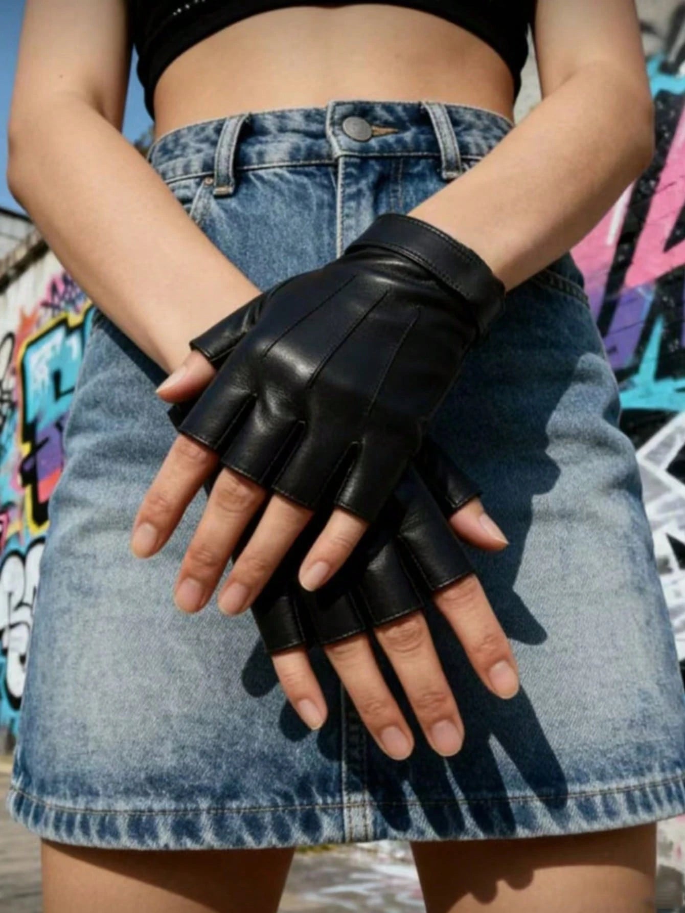 Black fingerless leather gloves worn by a model in a denim skirt, photographed outdoors against a graffiti wall.
