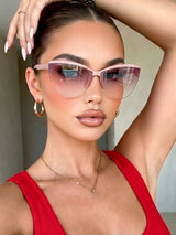 A close-up lifestyle shot of a woman wearing cat-eye sunglasses with a light pink frame and gradient tinted lenses, paired with a red ribbed top and gold jewelry.