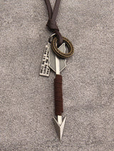 A decorative leather and metal pendant necklace featuring a brown leather cord, a silver-toned arrow-shaped charm, a small engraved metal tag, and a round bronze-toned medallion, laid flat on a gray textured surface.