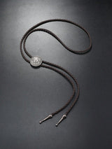 A dark brown braided bolo tie necklace with a silver-toned circular medallion and metal tips, displayed on a dark gray background.