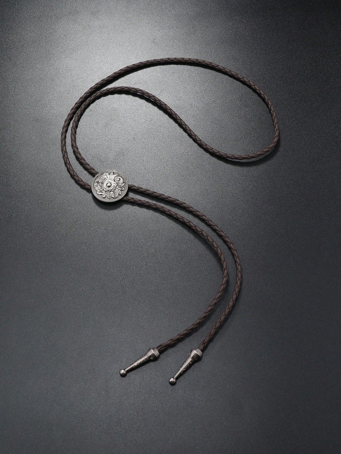 A dark brown braided bolo tie necklace with a silver-toned circular medallion and metal tips, displayed on a dark gray background.