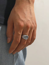 A close-up of a person's hand wearing a silver-toned ring with a stylized 'W' emblem on a textured band, paired with a dark shirt and light gray pants.