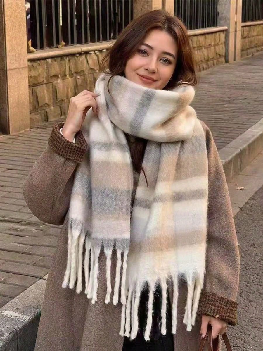 A woman wearing a thick, soft plaid scarf in beige, white, and gray tones, wrapped around her neck, paired with a brown coat, standing on a stone sidewalk.