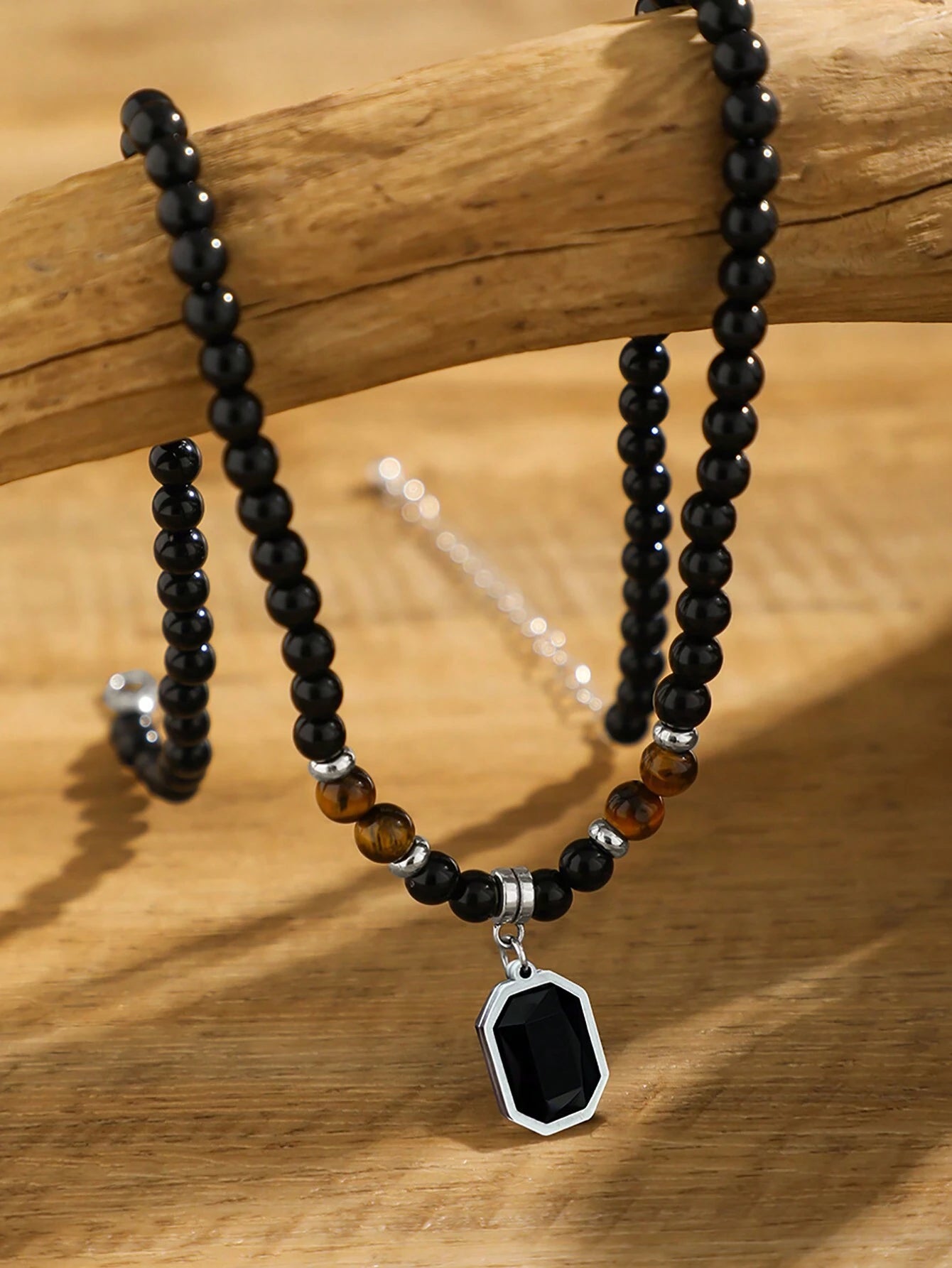A beaded necklace with black round beads, a few brown tiger-eye style beads, and a silver-toned pendant with a black faceted gemstone, displayed on a wooden branch over a wooden surface.