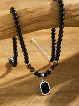 A beaded necklace with black round beads, a few brown tiger-eye style beads, and a silver-toned pendant with a black faceted gemstone, displayed on a wooden branch over a wooden surface.