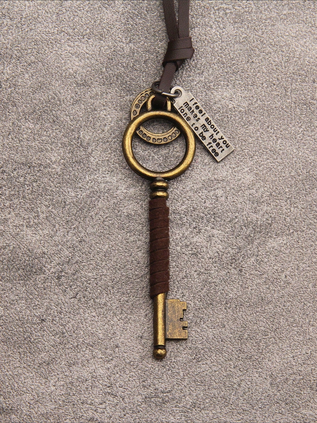 A vintage-style key-shaped pendant with a dark brown leather wrap, antique gold-toned metal key head and bit, and a small silver hanging tag, shown on a textured gray background.