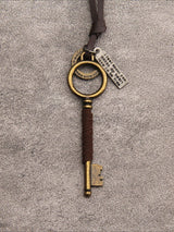 A vintage-style key-shaped pendant with a dark brown leather wrap, antique gold-toned metal key head and bit, and a small silver hanging tag, shown on a textured gray background.