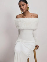 A woman models an off-the-shoulder white dress with a fluffy faux-fur collar, paired with chunky gold statement earrings, seated on a wooden stool against a plain light background.
