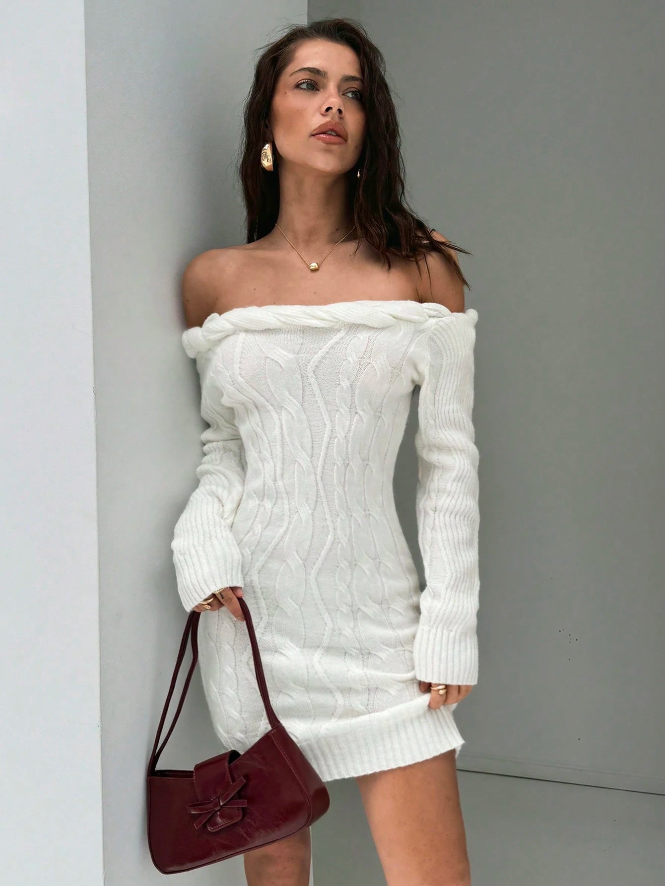 A woman models an off-the-shoulder white cable-knit mini dress, holding a burgundy shoulder bag against a minimalist gray wall.