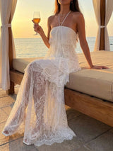A woman wearing a white lace halter maxi dress sits on a beach cabana daybed at sunset, holding a glass of white wine.