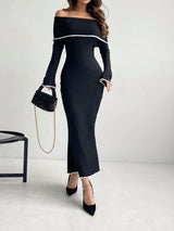 A woman models a black off-the-shoulder long-sleeve maxi dress with white trim details, paired with black pointed-toe heels and a black beaded handbag with a gold chain strap, set against a plain light gray studio background.