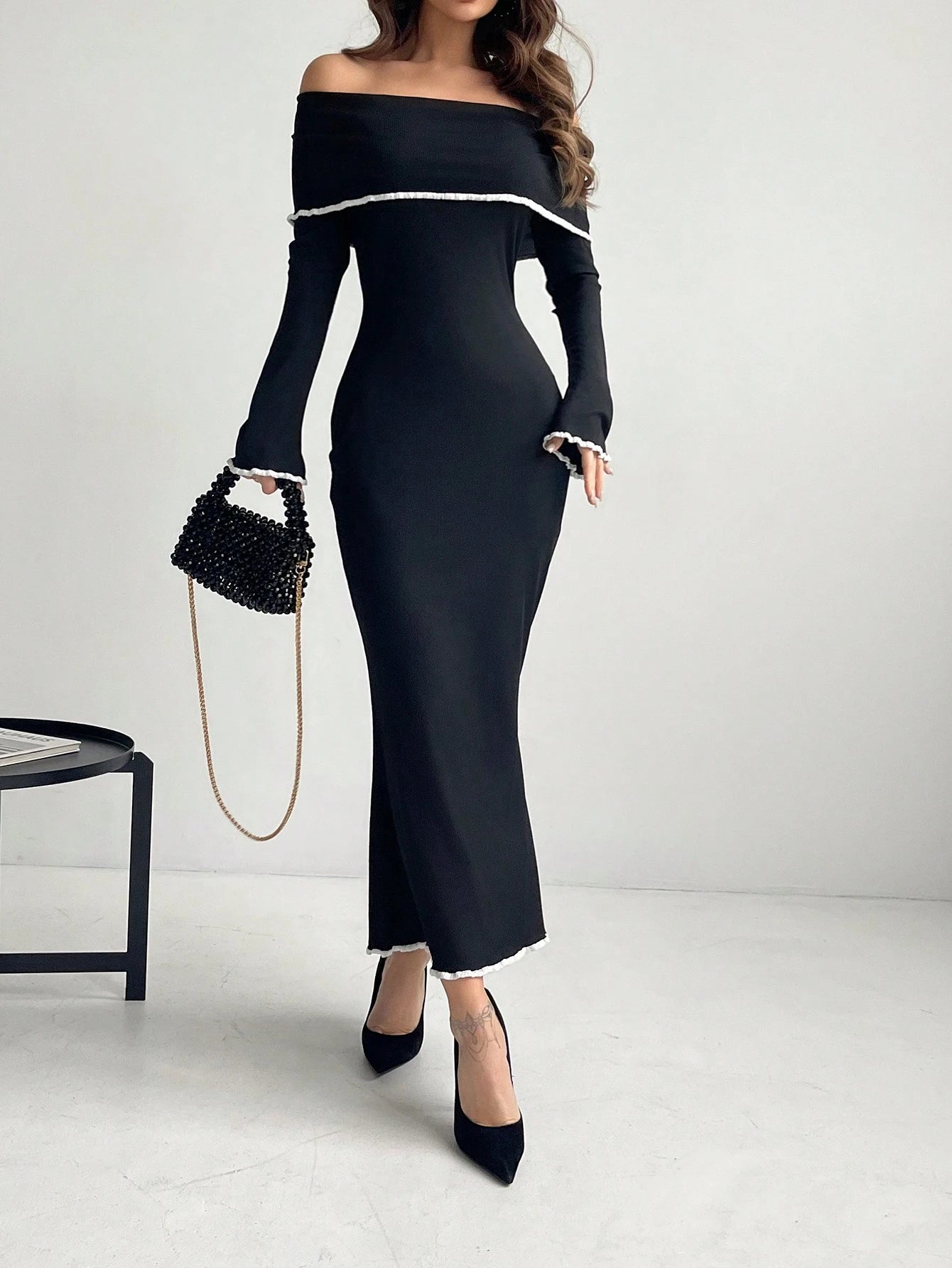 A woman models a black off-the-shoulder long-sleeve maxi dress with white trim details, paired with black pointed-toe heels and a black beaded handbag with a gold chain strap, set against a plain light gray studio background.