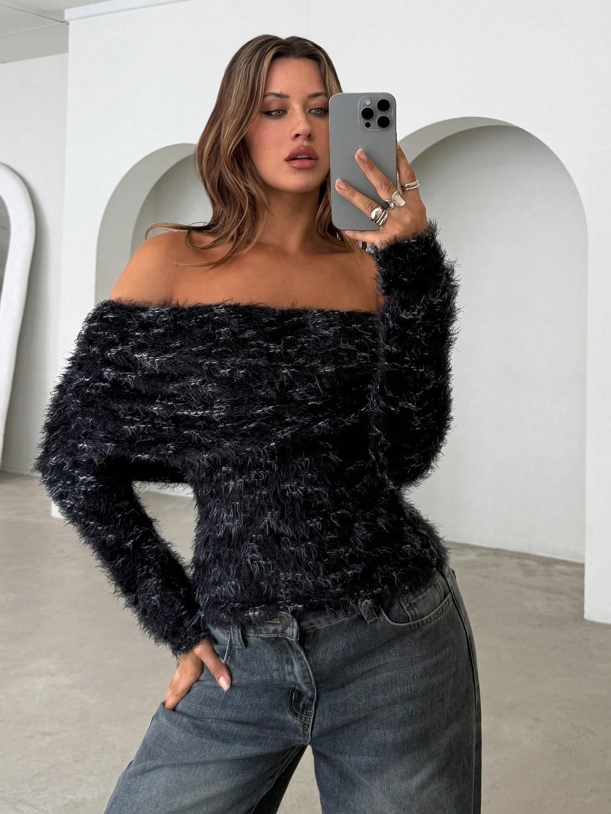 A woman takes a mirror selfie wearing a black and gray fuzzy off-the-shoulder long-sleeve sweater and dark gray jeans in a minimalist white room with arched walls.
