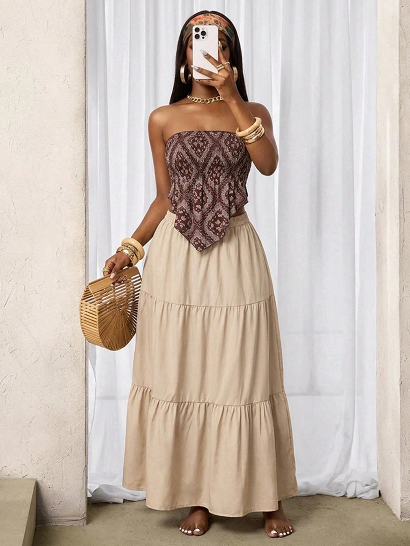Woman wearing a strapless top with intricate pattern and a long beige skirt, holding a woven bag indoors.