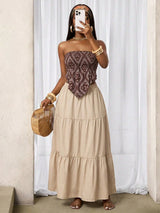 Woman wearing a strapless top with intricate pattern and a long beige skirt, holding a woven bag indoors.