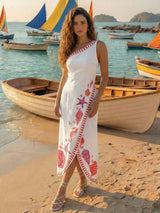 Woman in a white dress with red patterns standing on a beach with boats in the background