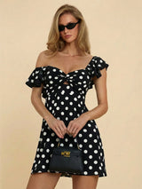 A woman modeling an off-the-shoulder black dress with white polka dots, featuring a sweetheart neckline with a front cutout and ruffled sleeves. She is holding a small black handbag with gold hardware against a solid beige background.