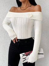 A woman wearing a ribbed off-the-shoulder white long-sleeve top with a floral embellishment on the chest, paired with black jeans and holding a magazine against a textured light gray wall.