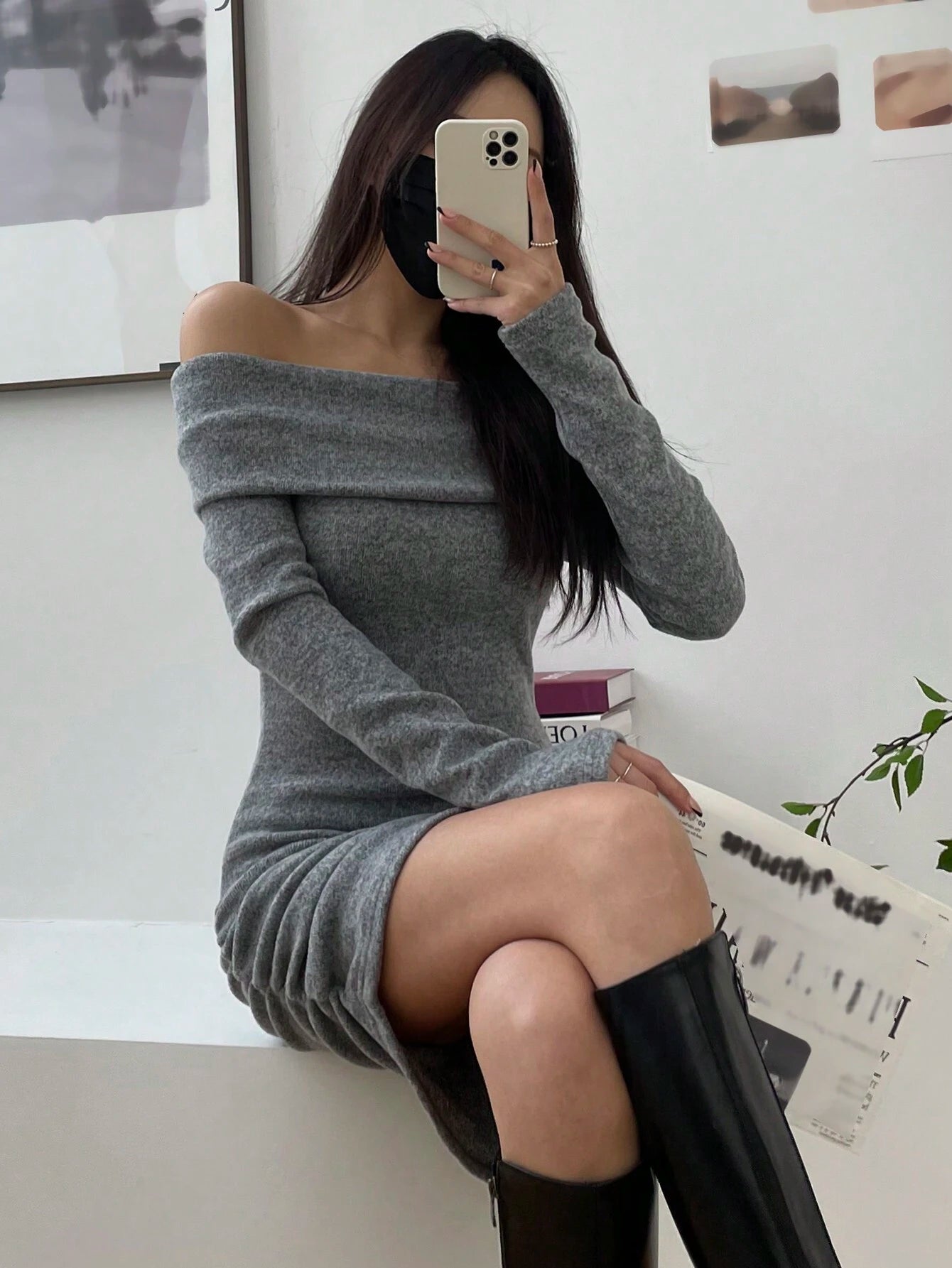 A woman takes a mirror selfie wearing a gray off-the-shoulder long-sleeve mini dress and black knee-high boots in a bright indoor setting.