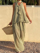 A woman wearing a sage green linen two-piece set consisting of a sleeveless ruffled top and matching wide-leg pants, holding a woven straw bag against a beige stucco wall background.