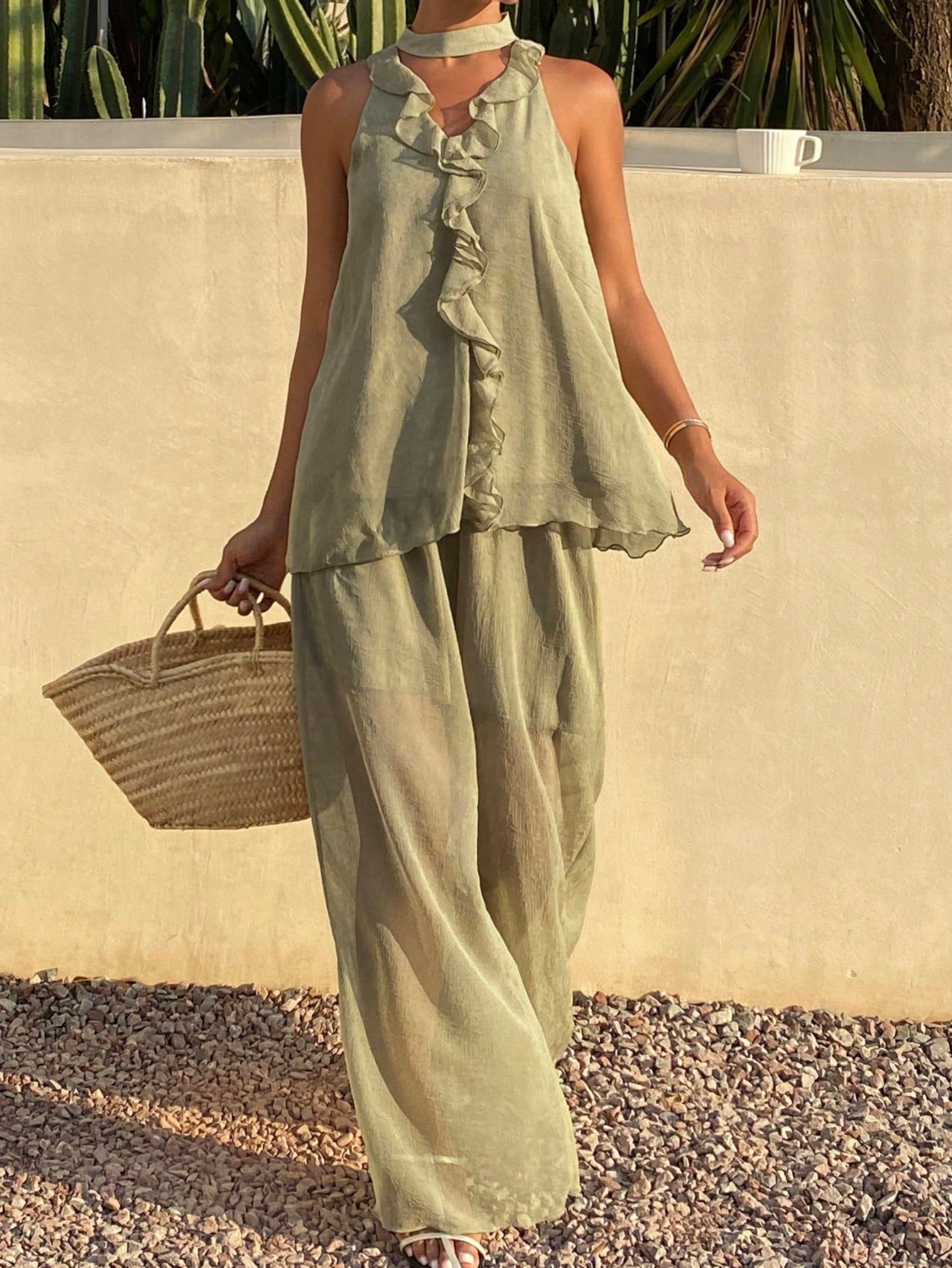 A woman wearing a sage green linen two-piece set consisting of a sleeveless ruffled top and matching wide-leg pants, holding a woven straw bag against a beige stucco wall background.