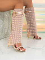 A close-up of a model's legs wearing sheer nude-beige thigh-high polka dot boot covers with metallic rose gold straps and gold heels, paired with matching metallic heeled sandals.