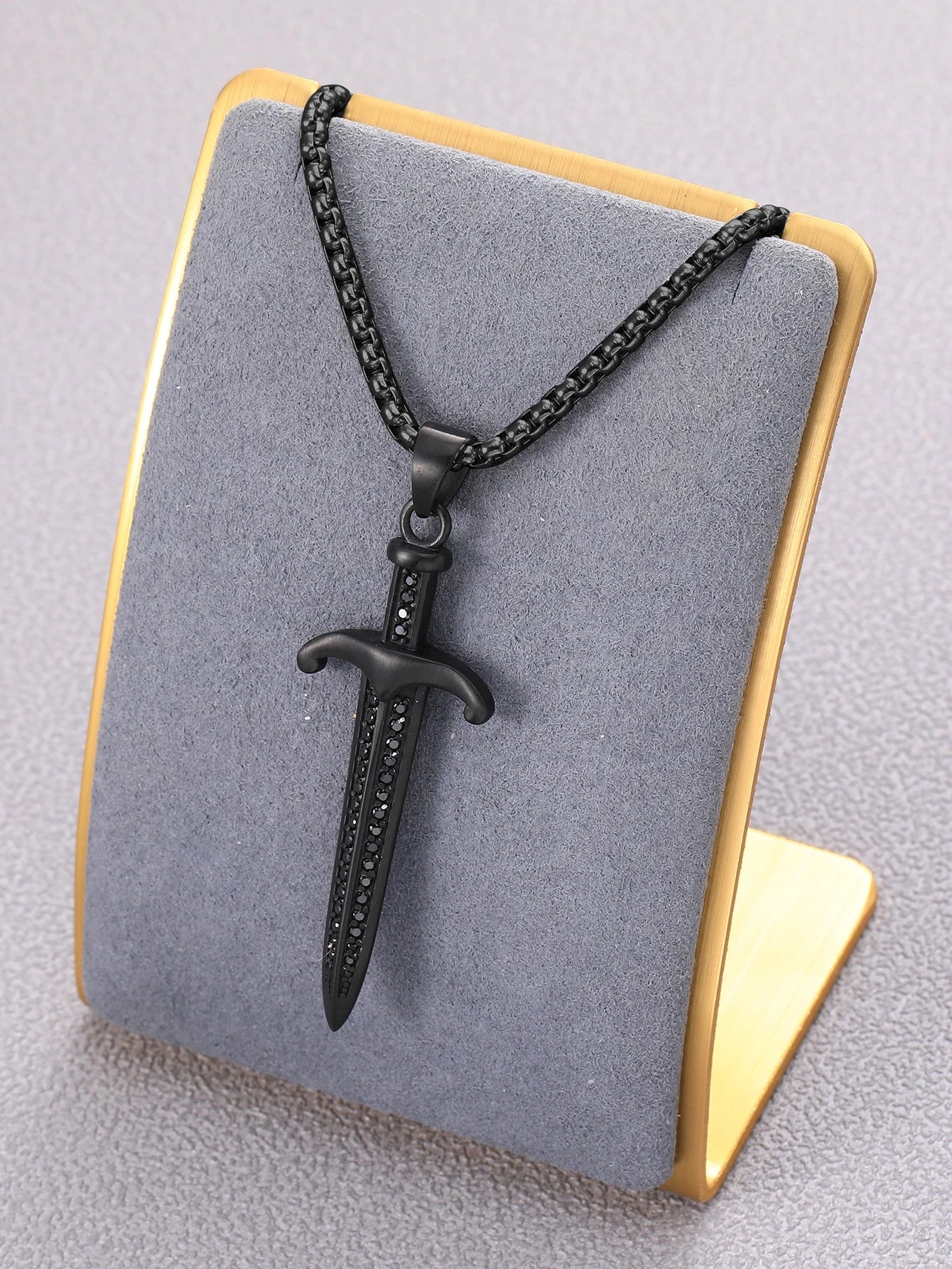 A black metal dagger pendant necklace displayed on a gray velvet jewelry bust with a gold-colored frame, set against a neutral gray background.