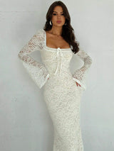 A woman modeling a fitted white lace maxi dress with long sheer bell sleeves, a square neckline, and a front tie detail, standing against a light gray wall.