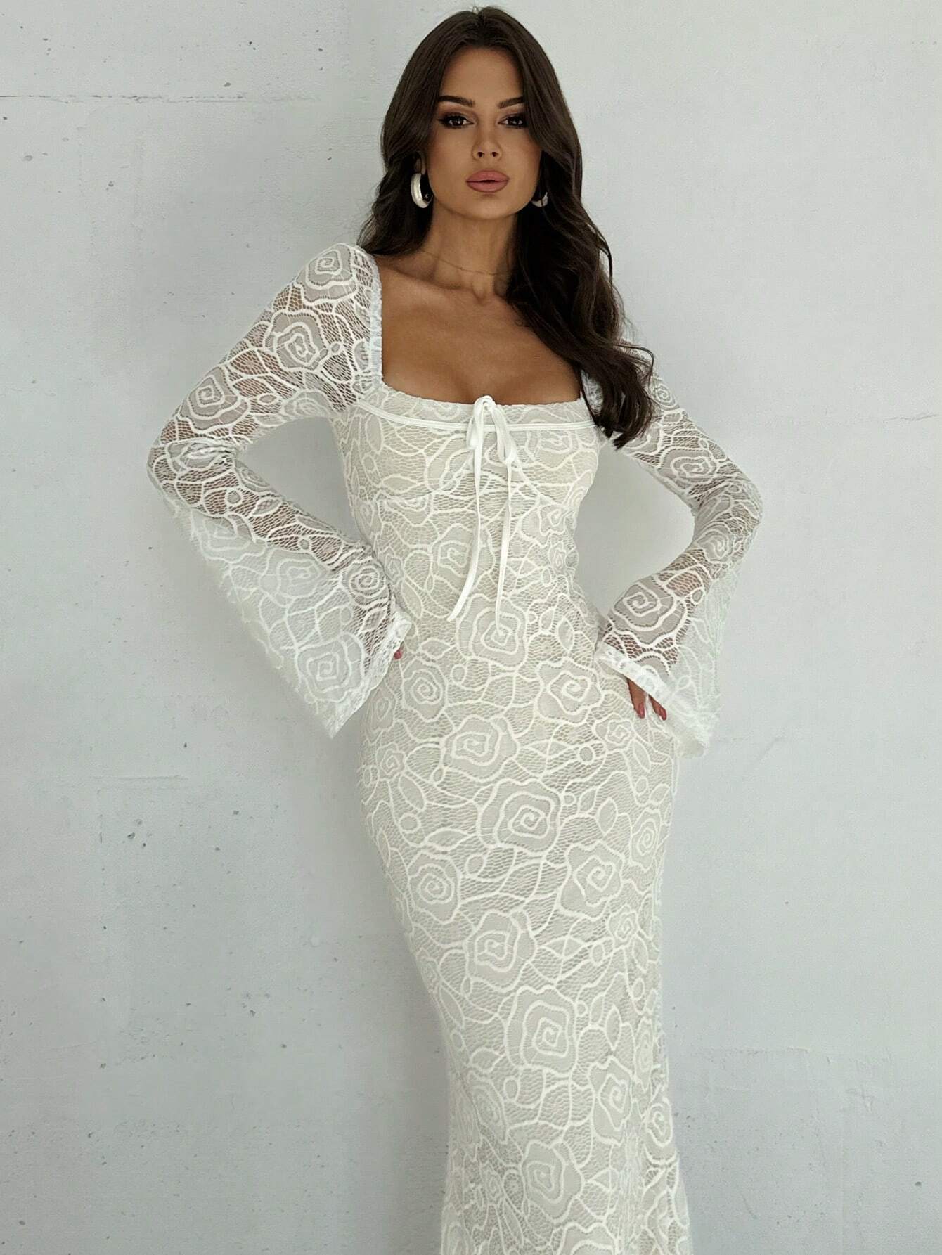 A woman modeling a fitted white lace maxi dress with long sheer bell sleeves, a square neckline, and a front tie detail, standing against a light gray wall.