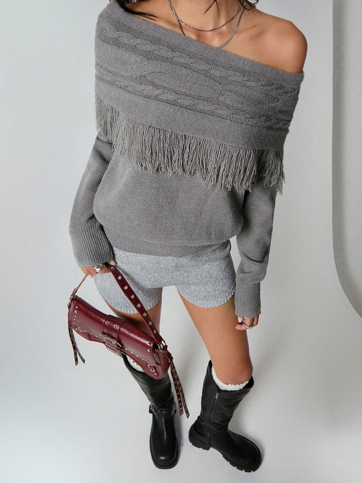 A model wearing a gray off-the-shoulder knit sweater with fringe trim, paired with a gray mini skirt, black knee-high boots, and holding a burgundy studded shoulder bag against a plain light gray background.