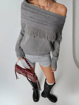 A model wearing a gray off-the-shoulder knit sweater with fringe trim, paired with a gray mini skirt, black knee-high boots, and holding a burgundy studded shoulder bag against a plain light gray background.