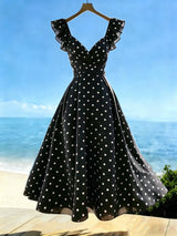 A black A-line dress with white polka dots and ruffled off-the-shoulder straps, hanging on a wooden hanger against a beach and blue sky background.