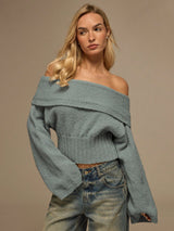 A blonde woman wearing a fuzzy off-the-shoulder cropped sweater in a dusty blue-gray color, paired with light-wash blue jeans, against a solid beige studio background.