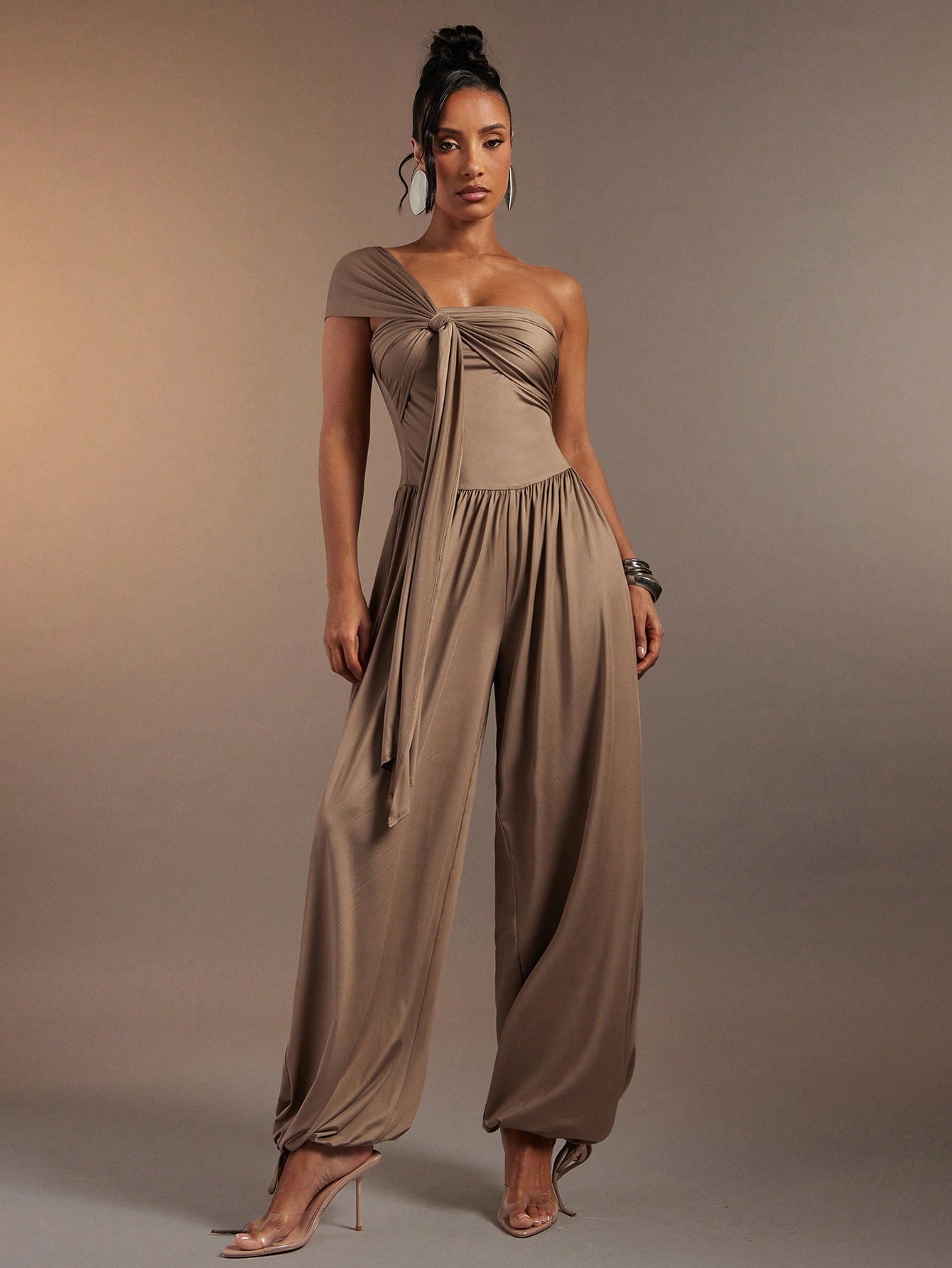 A woman modeling a taupe one-shoulder jumpsuit with a draped bodice and wide-leg pants, standing against a solid warm taupe studio background.