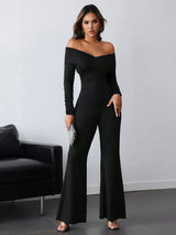 A woman modeling a black off-the-shoulder long-sleeve flared jumpsuit in a modern room setting with a black leather chair and a light gray wall.
