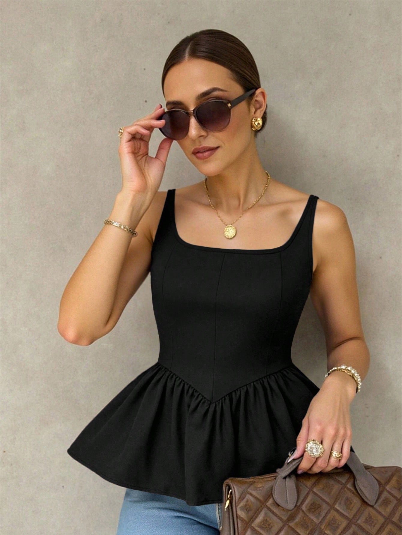 A woman wearing a black sleeveless peplum top, sunglasses, gold jewelry, and holding a brown quilted handbag against a textured gray wall.
