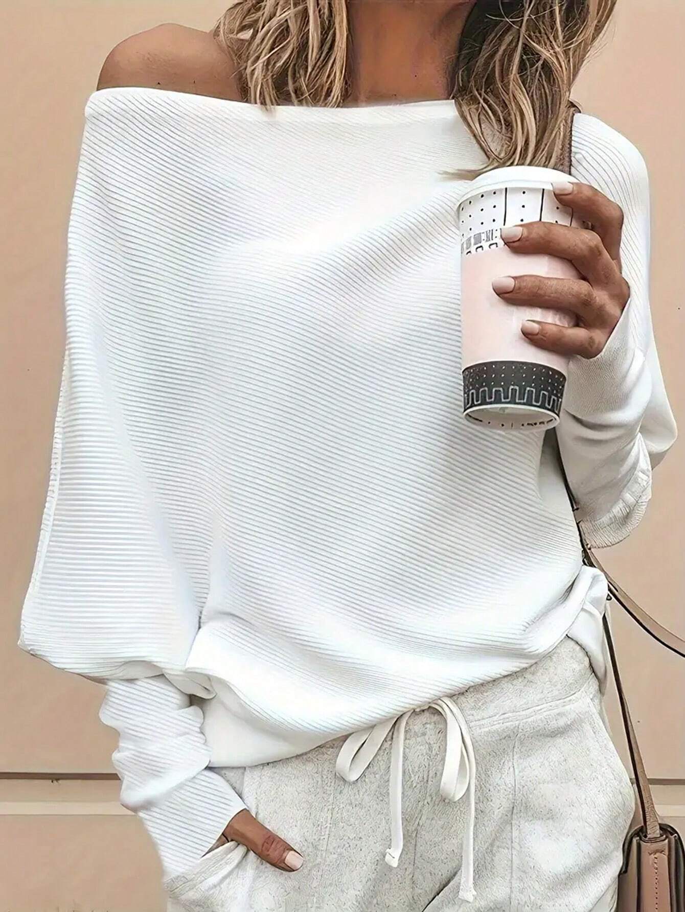A woman wearing a white off-the-shoulder ribbed knit sweater and light gray sweatpants, holding a light pink coffee cup against a beige wall.