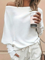 A woman wearing a white off-the-shoulder ribbed knit sweater and light gray sweatpants, holding a light pink coffee cup against a beige wall.
