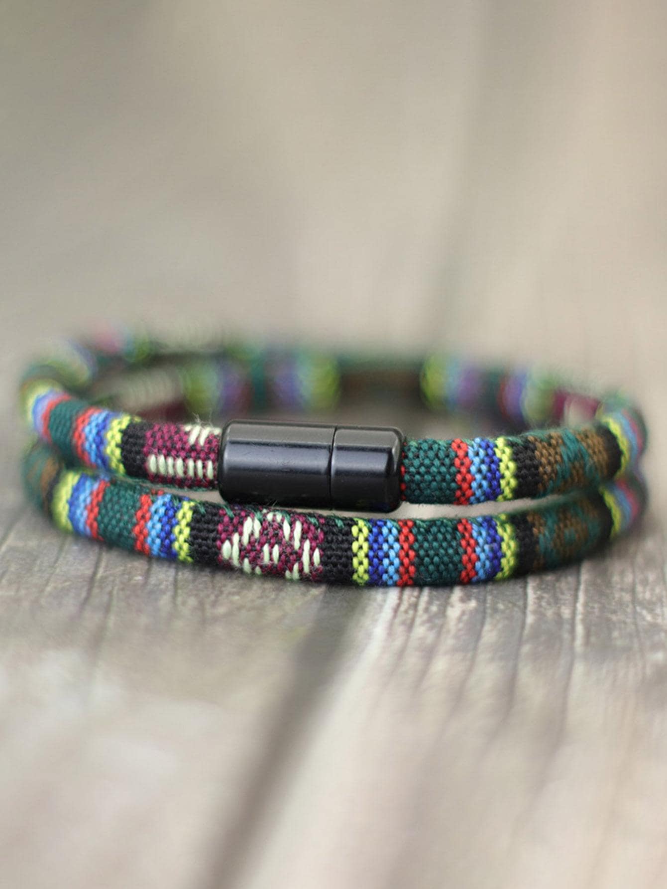 A close-up of a woven wrap bracelet with a black magnetic clasp, featuring a multicolor pattern of green, red, blue, yellow, and white threads on a wooden surface.