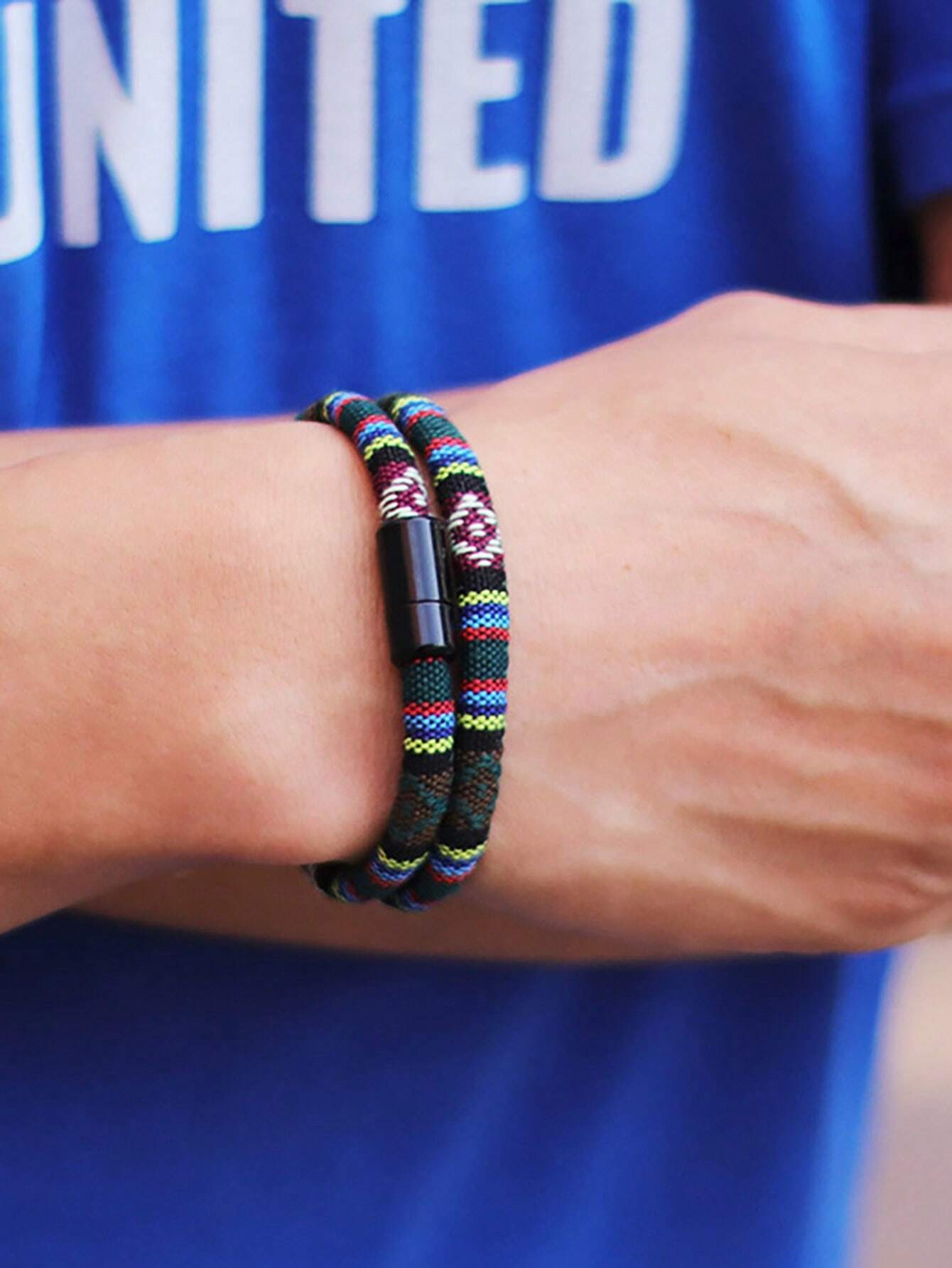 A close-up of a person's wrist wearing a multi-strand braided bracelet with a black magnetic clasp. The bracelet features black, green, red, yellow, and white woven cords, worn over a blue t-shirt.