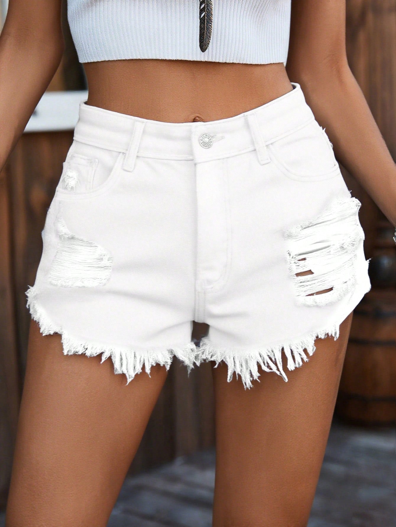 White distressed denim shorts with a raw frayed hem, worn by a model in front of a rustic wooden background.