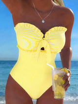 A woman wearing a bright yellow one-piece swimsuit with thin white vertical stripes, ruffled trim, and a front tie detail, standing on a beach with the ocean and blue sky in the background. She is holding a clear plastic drink cup with a straw.