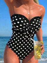 A woman wearing a black one-piece swimsuit with white polka dots and a bow detail at the bust, standing on a beach with the ocean and blue sky in the background while holding a drink.