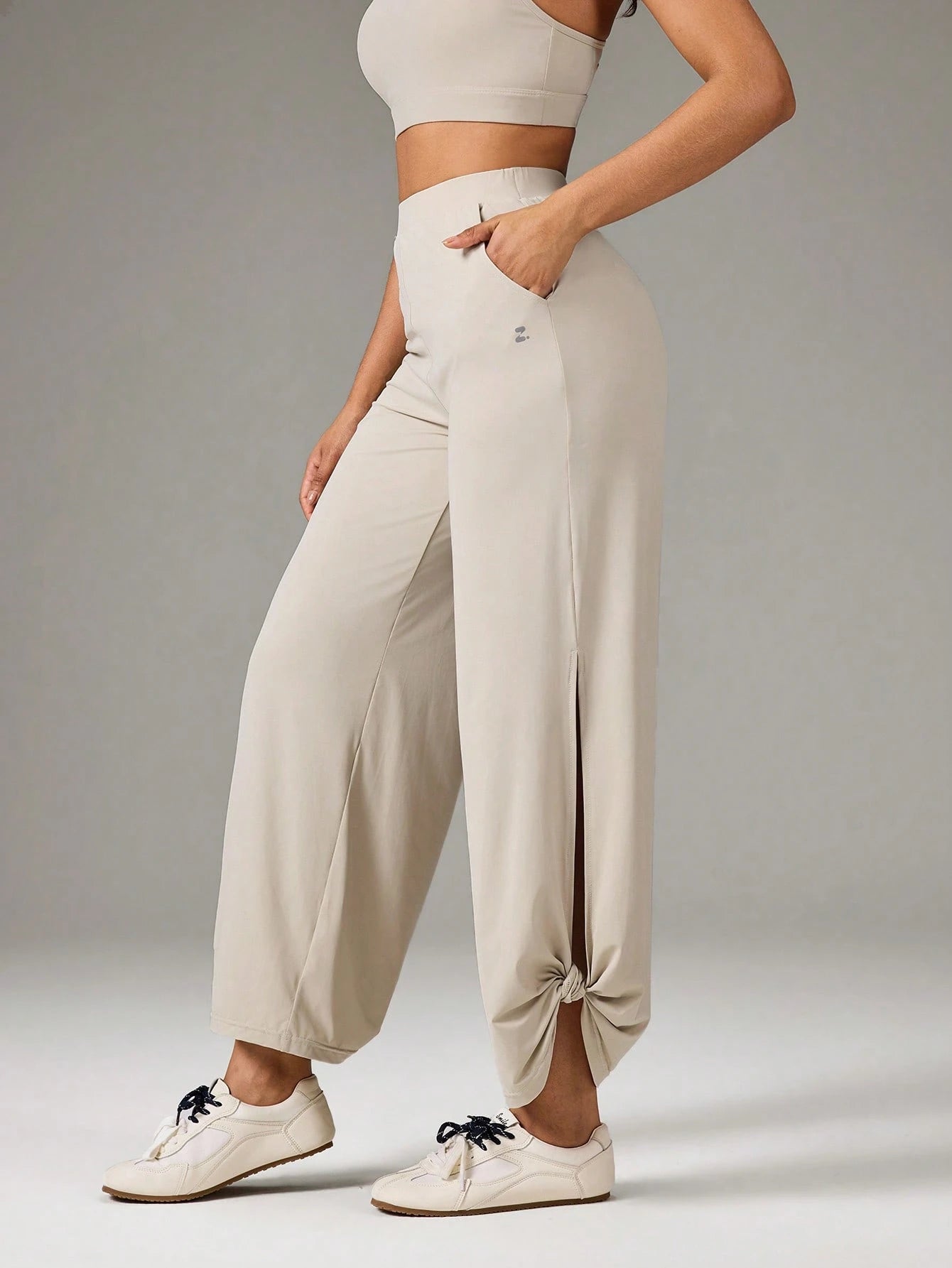A model wearing high-waisted, wide-leg jogger pants in a light beige color, styled with a matching beige sports bra and white sneakers against a neutral gray studio background.