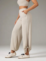 A model wearing high-waisted, wide-leg jogger pants in a light beige color, styled with a matching beige sports bra and white sneakers against a neutral gray studio background.