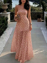 A woman wearing a strapless dusty pink maxi dress with white polka dots, holding a woven clutch on a stone pathway with a garden and water in the background.
