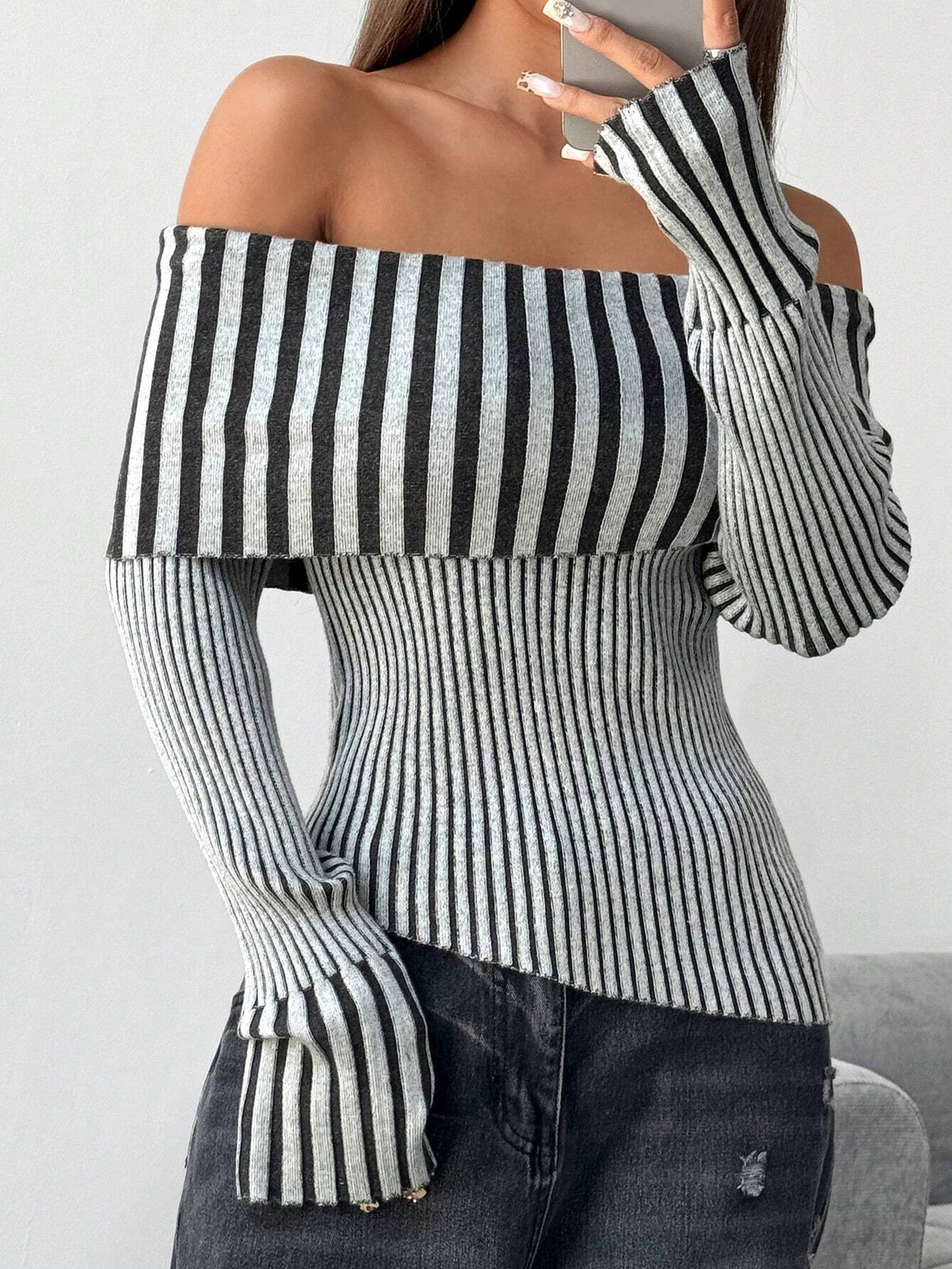 A woman wearing a black and white vertically striped off-the-shoulder long-sleeve knit top paired with dark gray jeans, shown in a close-up shot.
