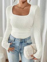 A woman wearing a fitted, ribbed long-sleeve sweater with a square neckline in off-white, paired with light-wash blue jeans and holding a matching off-white quilted clutch bag.