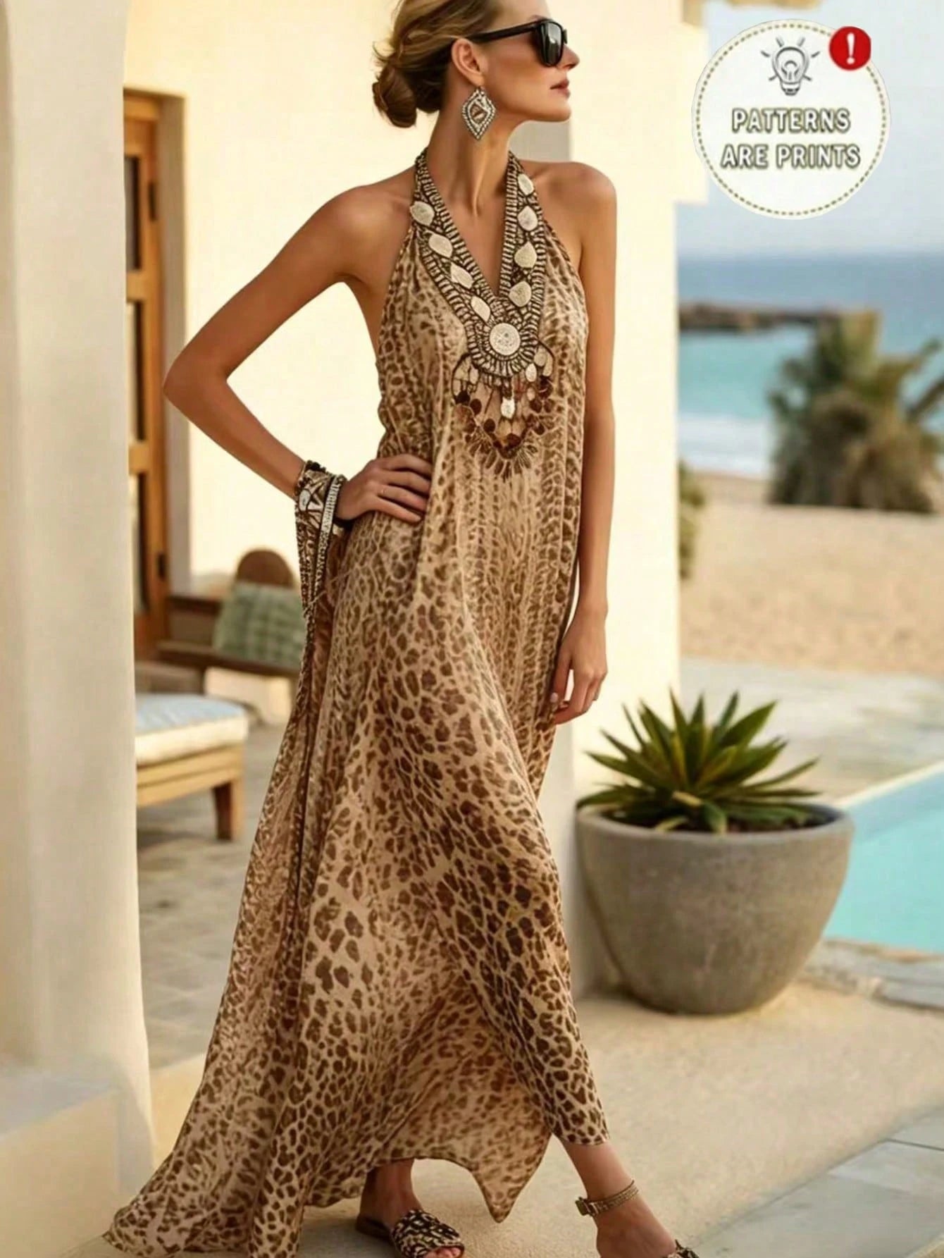 A woman models a sleeveless halter maxi dress with a beige and brown leopard print pattern, styled with a chunky statement necklace and sunglasses in an outdoor patio setting with a beach background.